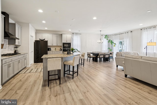 kitchen with wall chimney range hood, hanging light fixtures, light stone counters, black appliances, and a center island with sink