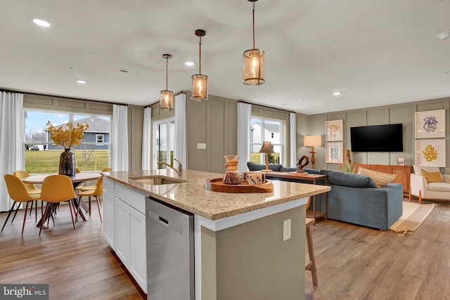 kitchen featuring light stone counters, stainless steel dishwasher, a kitchen island with sink, sink, and hanging light fixtures