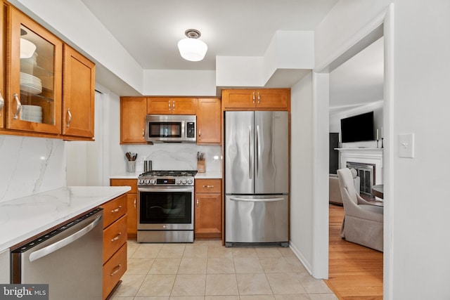 kitchen featuring stainless steel appliances, brown cabinetry, glass insert cabinets, and backsplash