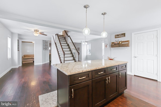 kitchen with light stone counters, dark brown cabinets, ceiling fan, dark wood-type flooring, and pendant lighting