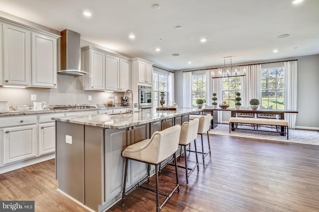 kitchen with a center island with sink, wall chimney exhaust hood, decorative light fixtures, a kitchen bar, and stainless steel appliances