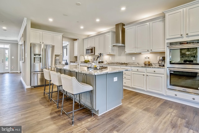 kitchen with white cabinets, appliances with stainless steel finishes, a kitchen island with sink, and wall chimney exhaust hood