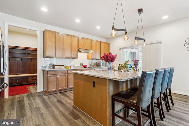 kitchen with decorative backsplash, a breakfast bar area, light stone counters, dark wood-style flooring, and under cabinet range hood