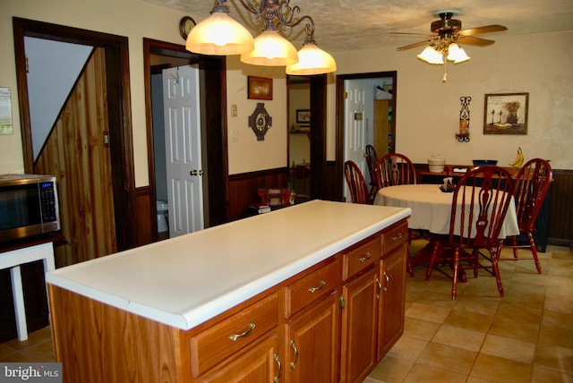 kitchen with light tile patterned flooring, wooden walls, a kitchen island, pendant lighting, and ceiling fan with notable chandelier