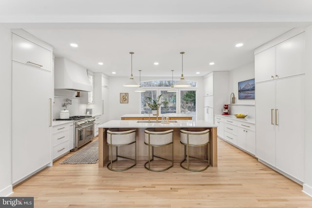 kitchen featuring white cabinetry, an island with sink, custom range hood, light hardwood / wood-style floors, and range with two ovens