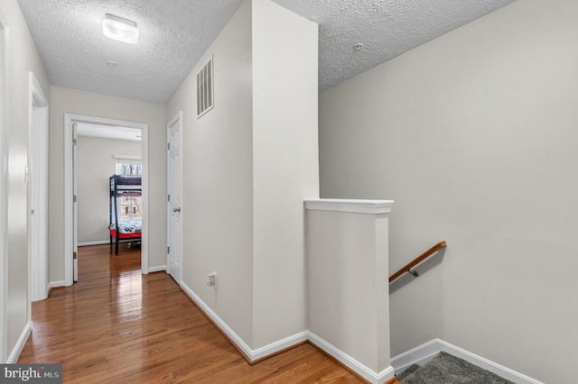 hall featuring visible vents, light wood finished floors, baseboards, a textured ceiling, and an upstairs landing