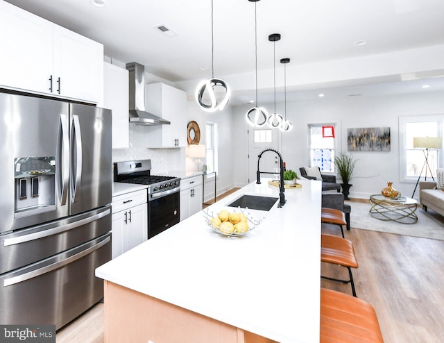 kitchen with stainless steel appliances, wall chimney range hood, a kitchen island with sink, and white cabinetry