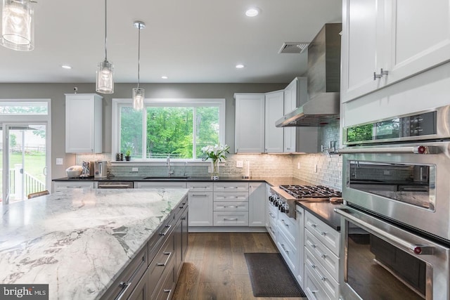 kitchen with wall chimney range hood, hanging light fixtures, appliances with stainless steel finishes, white cabinets, and dark stone counters