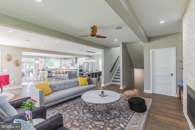 living room with ceiling fan, dark hardwood / wood-style floors, a stone fireplace, and beamed ceiling