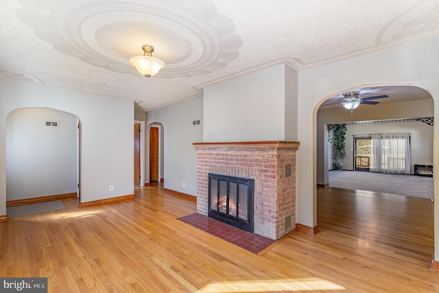 unfurnished living room featuring arched walkways, a textured ceiling, wood finished floors, a ceiling fan, and a brick fireplace