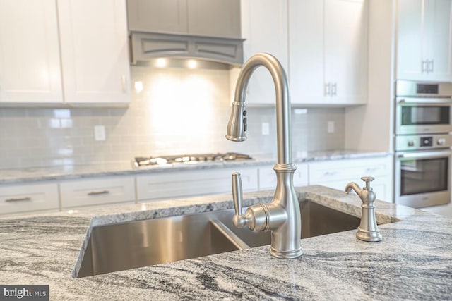 kitchen featuring double oven, light stone counters, a sink, white cabinetry, and tasteful backsplash