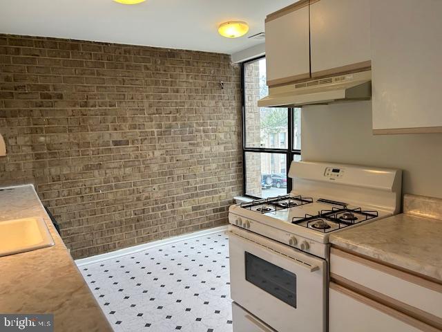 kitchen featuring brick wall, under cabinet range hood, light countertops, and white range with gas stovetop