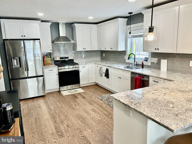 kitchen featuring wall chimney range hood, white cabinetry, hanging light fixtures, stainless steel appliances, and light hardwood / wood-style floors