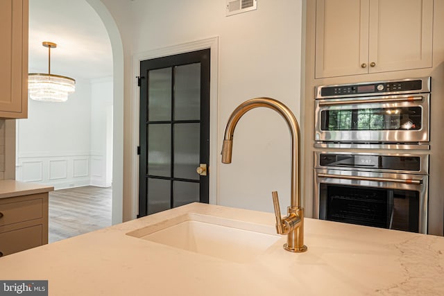 kitchen with light stone countertops, stainless steel double oven, sink, hanging light fixtures, and a notable chandelier