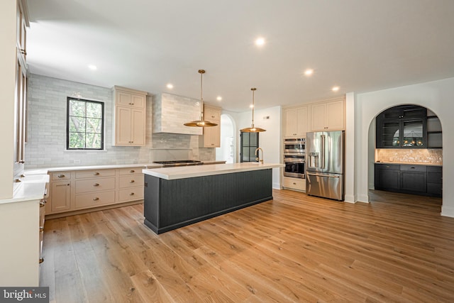 kitchen with wall chimney range hood, stainless steel appliances, light hardwood / wood-style floors, hanging light fixtures, and a center island with sink