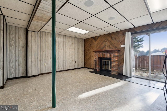 unfurnished living room featuring a brick fireplace, a drop ceiling, wooden walls, and baseboards