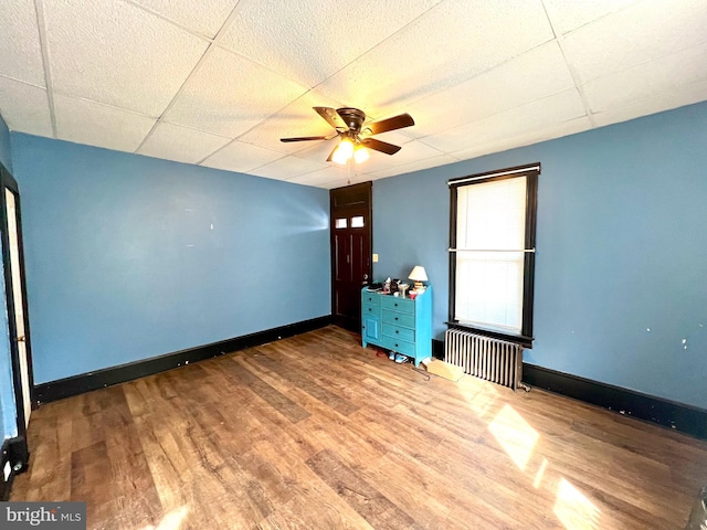 empty room with a drop ceiling, hardwood / wood-style flooring, radiator heating unit, and ceiling fan