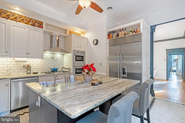 kitchen with stainless steel appliances, an island with sink, wall chimney range hood, and tasteful backsplash