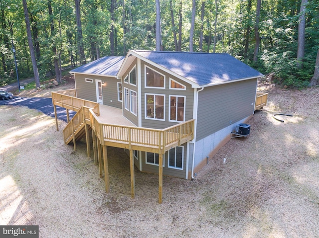 view of front facade with central AC, a shingled roof, dirt driveway, a wooden deck, and a forest view