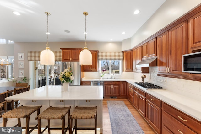 kitchen featuring decorative backsplash, under cabinet range hood, appliances with stainless steel finishes, a kitchen breakfast bar, and brown cabinets