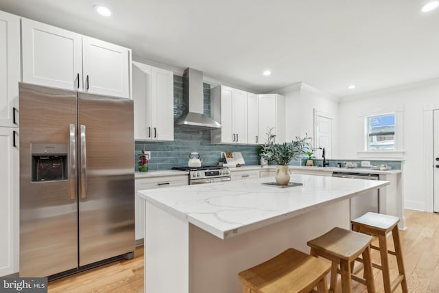 kitchen featuring wall chimney range hood, a kitchen island, a kitchen bar, stainless steel appliances, and white cabinets