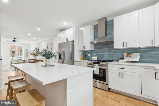 kitchen featuring white cabinets, appliances with stainless steel finishes, wall chimney exhaust hood, backsplash, and a breakfast bar area