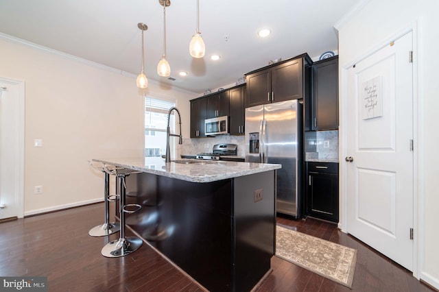 kitchen with appliances with stainless steel finishes, pendant lighting, tasteful backsplash, light stone counters, and a center island with sink