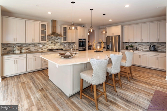 kitchen with a kitchen island with sink, wall chimney range hood, decorative backsplash, appliances with stainless steel finishes, and white cabinetry