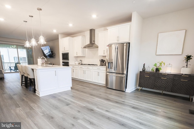 kitchen featuring wall chimney range hood, hanging light fixtures, black appliances, an island with sink, and white cabinets