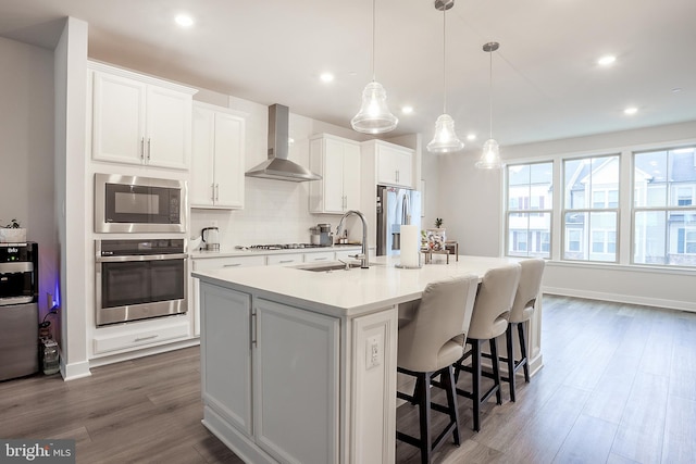 kitchen with wall chimney exhaust hood, appliances with stainless steel finishes, a kitchen island with sink, and pendant lighting