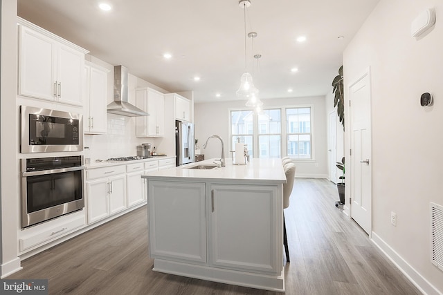 kitchen featuring wall chimney exhaust hood, sink, an island with sink, stainless steel appliances, and white cabinets