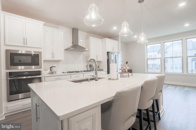 kitchen with appliances with stainless steel finishes, pendant lighting, sink, wall chimney range hood, and a center island with sink