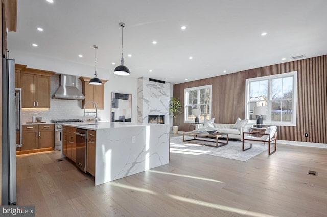 kitchen with a center island with sink, stainless steel appliances, pendant lighting, light stone counters, and wall chimney exhaust hood