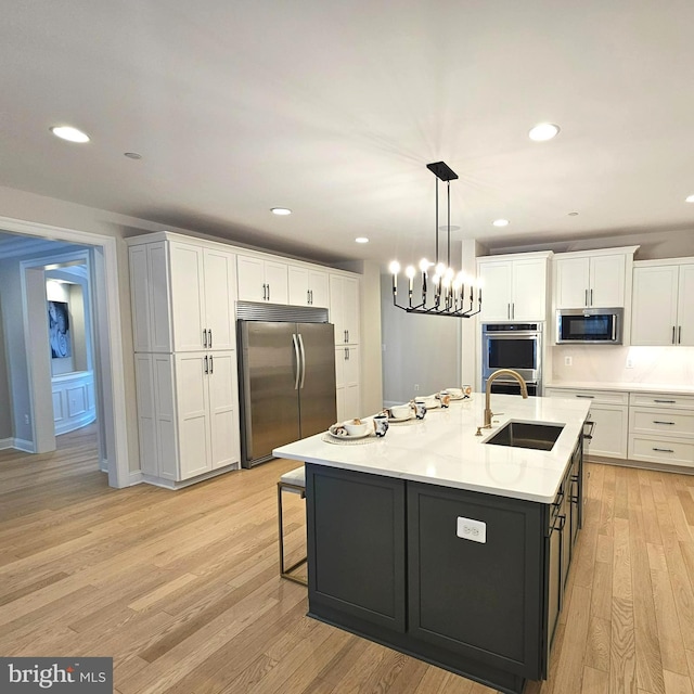 kitchen featuring white cabinetry, stainless steel appliances, an island with sink, sink, and a notable chandelier