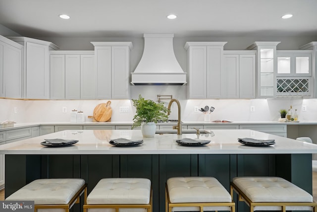 kitchen featuring backsplash, a center island with sink, custom exhaust hood, a kitchen bar, and white cabinets