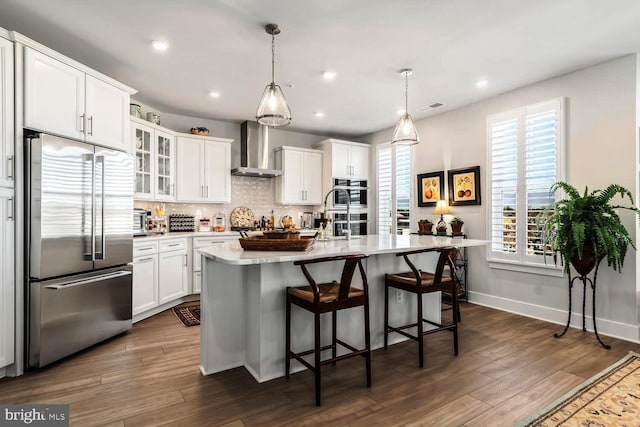 kitchen with appliances with stainless steel finishes, hanging light fixtures, an island with sink, white cabinets, and wall chimney range hood