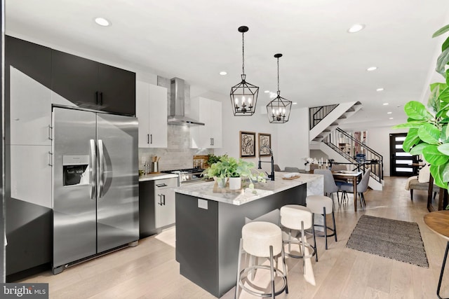 kitchen with a center island with sink, stainless steel fridge, wall chimney range hood, decorative light fixtures, and tasteful backsplash