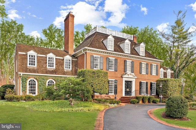 colonial house with aphalt driveway, brick siding, roof with shingles, a chimney, and a front yard