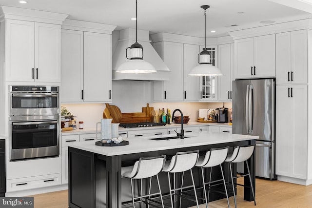 kitchen featuring premium range hood, decorative light fixtures, an island with sink, sink, and stainless steel appliances