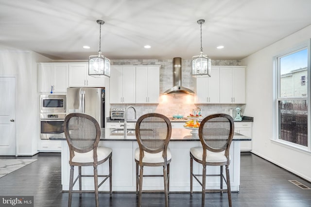 kitchen with appliances with stainless steel finishes, white cabinetry, a center island with sink, and wall chimney range hood