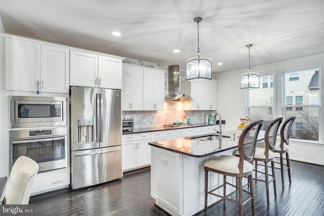 kitchen featuring a kitchen island with sink, stainless steel appliances, a sink, wall chimney range hood, and dark countertops