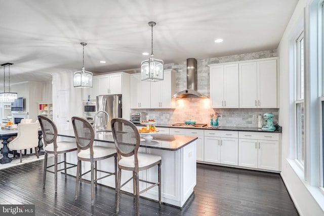 kitchen featuring white cabinetry, hanging light fixtures, wall chimney range hood, dark countertops, and a center island with sink