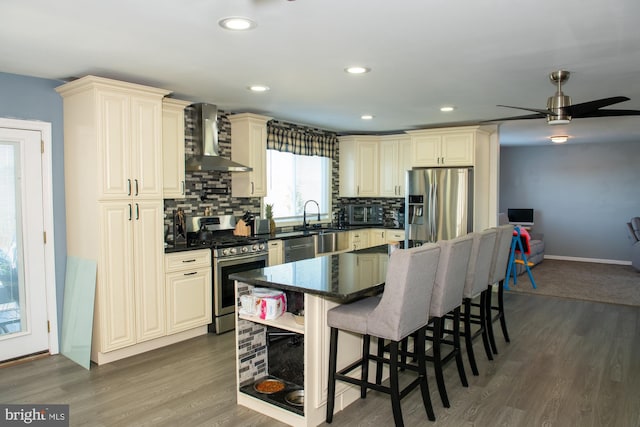 kitchen with stainless steel appliances, a kitchen island, wall chimney range hood, cream cabinetry, and tasteful backsplash