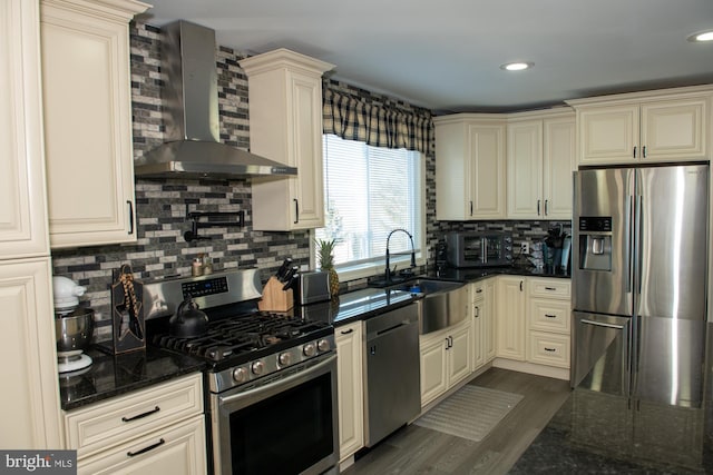 kitchen with cream cabinets, stainless steel appliances, a sink, wall chimney range hood, and dark stone counters