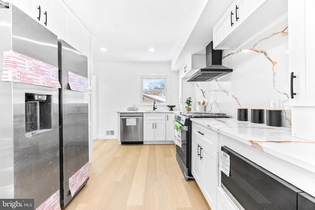 kitchen with light stone counters, stainless steel appliances, wall chimney range hood, white cabinets, and light hardwood / wood-style floors