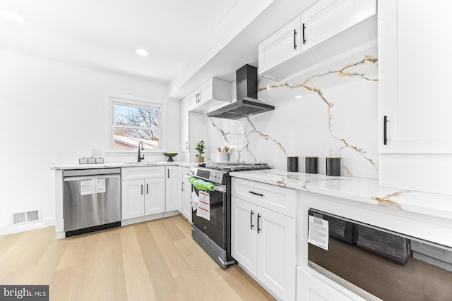 kitchen with wall chimney range hood, sink, light stone countertops, appliances with stainless steel finishes, and white cabinetry