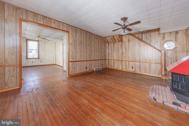 unfurnished living room featuring ceiling fan, wooden walls, a wood stove, and hardwood / wood-style flooring