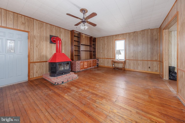 unfurnished living room featuring ceiling fan, hardwood / wood-style floors, wood walls, and a wood stove