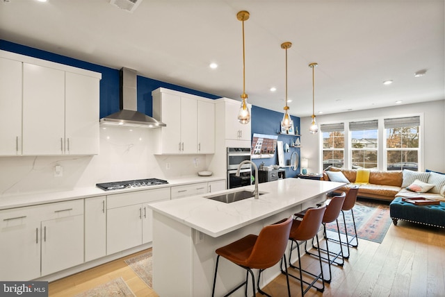 kitchen with white cabinets, appliances with stainless steel finishes, wall chimney exhaust hood, and an island with sink