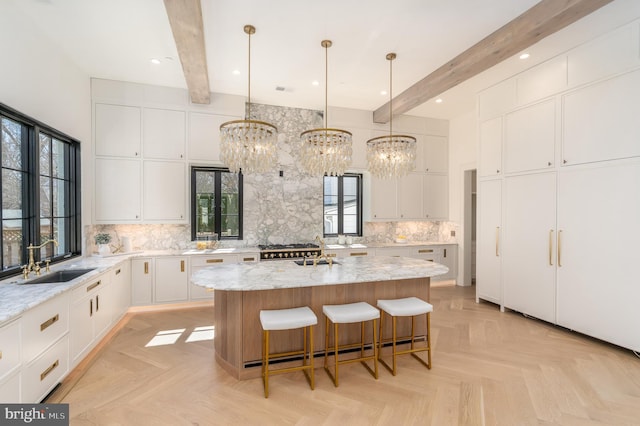 kitchen featuring light stone counters, a kitchen island with sink, a sink, beamed ceiling, and backsplash